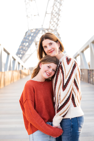 a mom and teen daughter stand together holding hands and hugging wearing sweaters on the Skydance Bridge at Scissortail Park in Oklahoma City