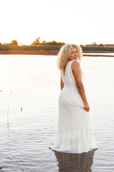 senior girl with curly blond hair wearing a long white dress stands ankle deep in Lake Hefner and looks over her shoulder with the sunset behind her