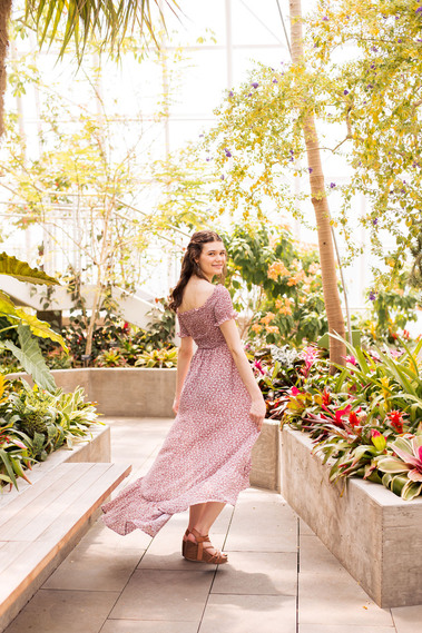 senior girl with brown hair wearing a muted red print dress twirls on a path and looks over her shoulder smiling at Crystal Bridge Conservatory in Myriad Gardens Oklahoma City