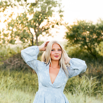 high school senior girl with blond hair wearing a light blue romper stands with her hands in her hair smiling softly in a field with green grass and trees in central Oklahoma