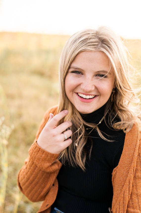 portrait of a high school senior girl with blond hair in a black turtleneck and rust colored cardigan, smiling and holding her hair as the wind blows