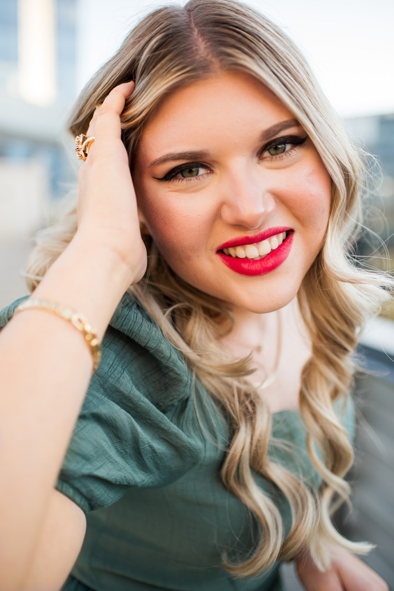 close up portrait of a blond high school senior girl with pink lipstick smiling with her hand in her hair wearing gold jewelry on the Devon Tower parking garage rooftop in downtown Oklahoma City