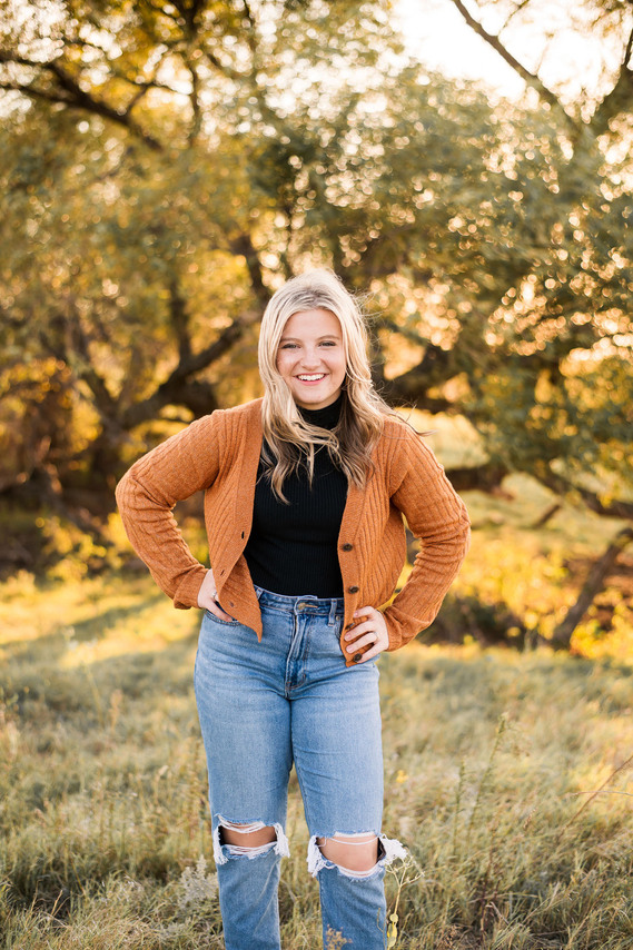 blond high school senior girl wearing jeans and an orange cardigan smiles with her hands on her hips with fall trees behind her in a field in Tuttle, Oklahoma