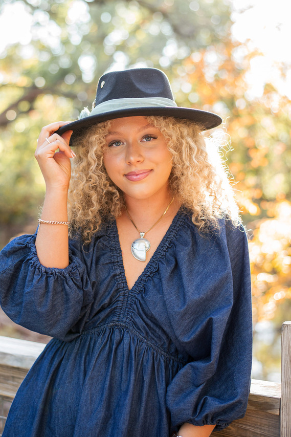 senior girl wearing a blue denim dress and wide brim hat leans against a bridge with one hand on brim softly smiling with fall trees behind her at Martin Nature Park in OKC
