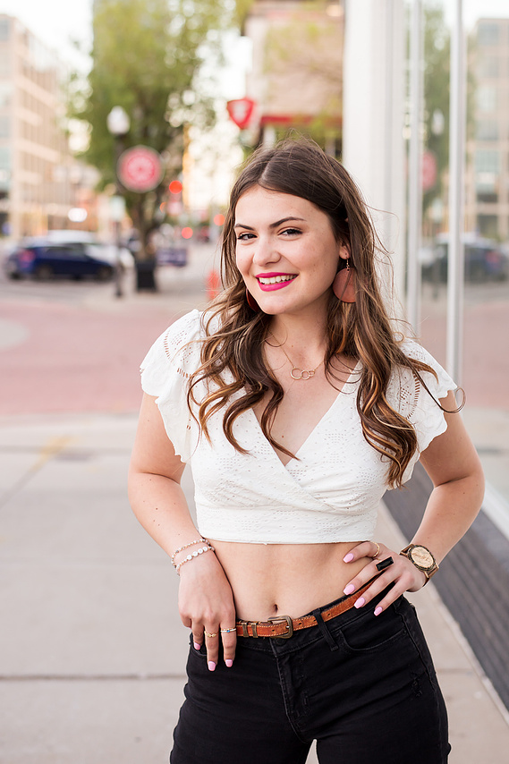 senior girl with long brown hair wearing a white cropped top and black jeans stands with hands on hips and smiles on a sidewalk  in Automobile Alley in Oklahoma City