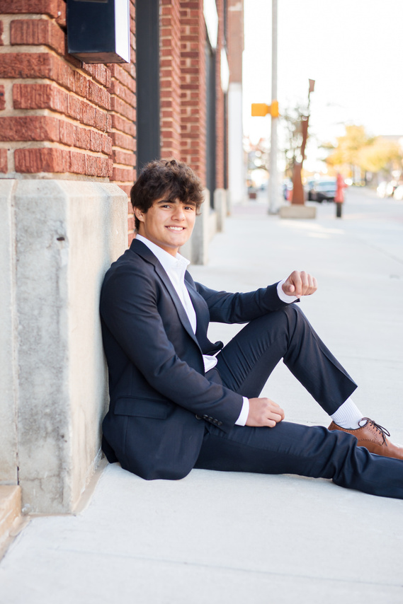 Senior guy wearing a dark suit and white dress shirt sits against a brick wall with one knee up and smiling. senior portrait outfits for guys