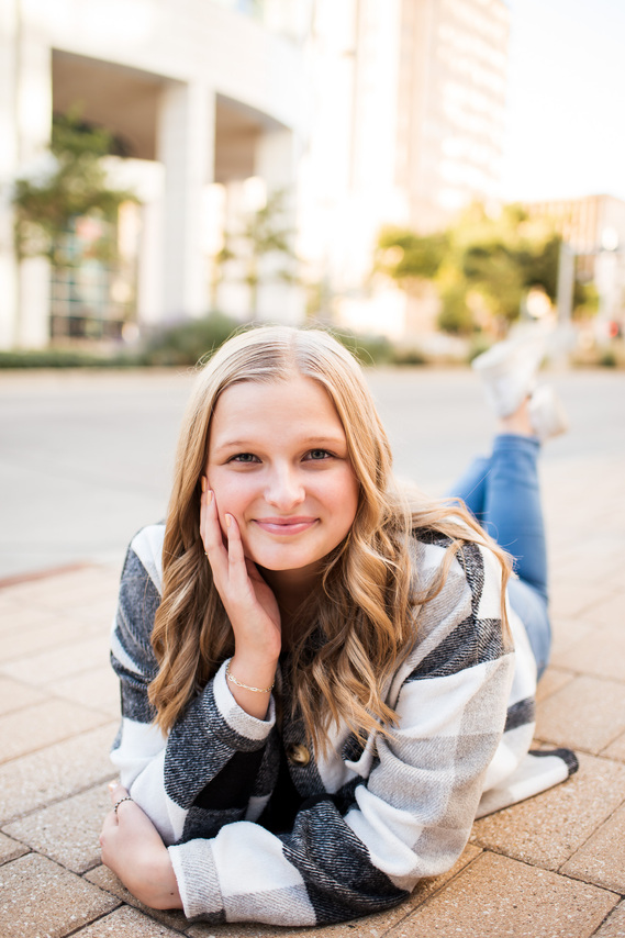 blond high school senior girl wearing a plaid jacket and jeans lays on her stomach on a sidewalk with one hand on her chin and smiles softly in downtown Oklahoma City