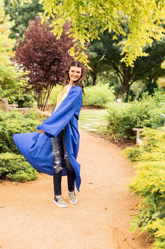 senior girl wearing a blue graduation gown twirls and smiles playfully standing on a path at the Myriad Gardens in OKC with trees and plants around her
