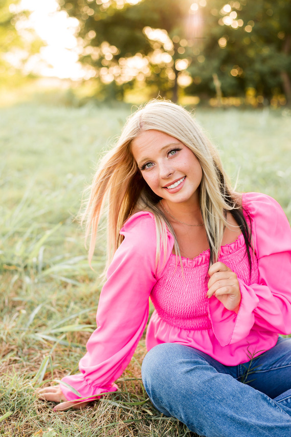 blond high school senior girl wearing a pink top and jeans sits in a field and smiles with one hand holding the end of her hair in Tuttle Oklahoma