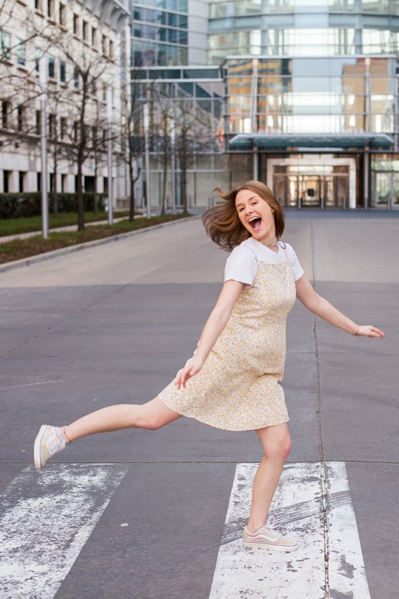 high school senior girl wearing a yellow dress and sneakers leaps along a crosswalk laughing with her arms out on a street in front of the Devon tower in Oklahoma City