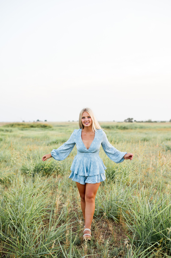blond high school senior girl wearing a blue romper walks forward with her arms outstretched and smiles in a field of tall grass in Tuttle, Oklahoma