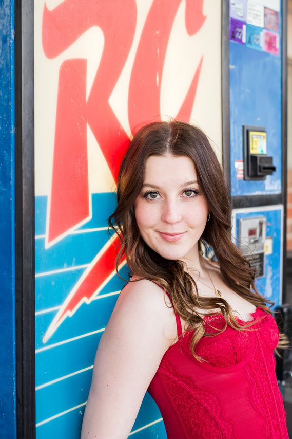 brunette high school senior girl wearing a red lacy camisole leans against a vintage RC Cola vending machine and smiles at camera in Tuttle, Oklahoma