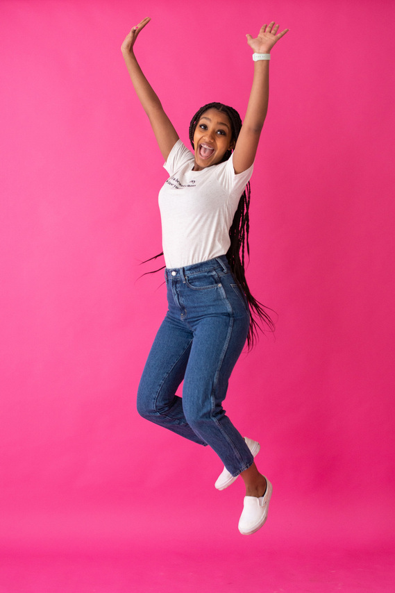 young woman of color wearing a white graphic tee and blue jeans jumps joyfully in the air with her arms up smiling in front of a hot pink seamless backdrop at a brand photo shoot in Oklahoma City