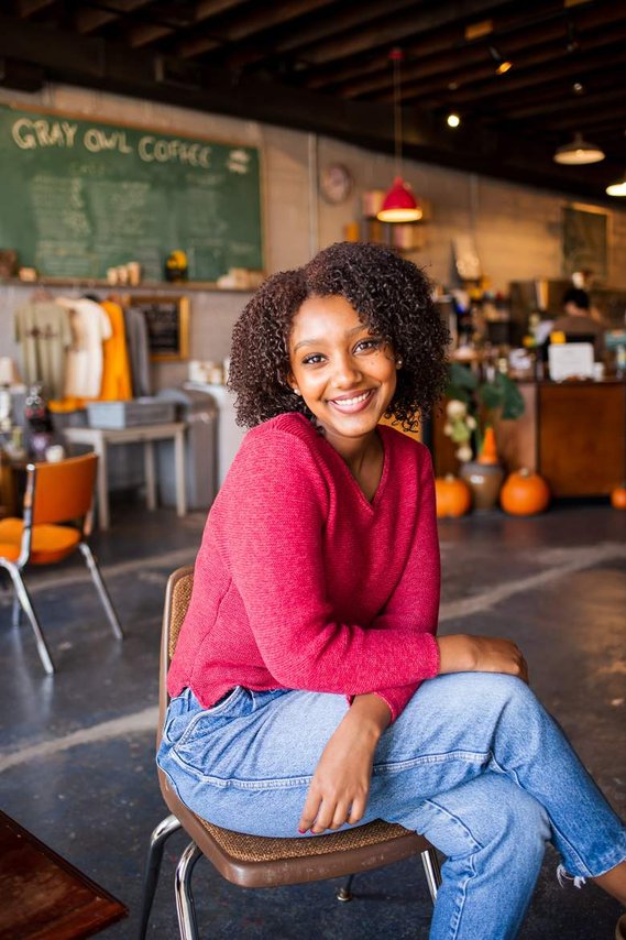 senior portrait of a black girl with curly hair wearing a pink sweater and blue jeans sitting cross legged and smiling inside a coffee shop in Norman Oklahoma with a coffee chalkboard menu behind her
