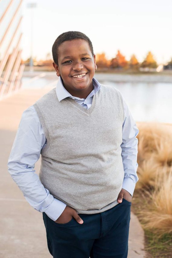 Teen Ethiopian boy in a blue dress shirt and gray sweater stands with hands in pockets and smiles by the Devon Boathouse in Oklahoma City