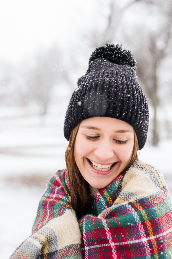 high school senior girl wrapped in a tartan scarf wearing a black beanie laughs over her shoulder standing in the snow in Schrock Park in Tuttle Oklahoma