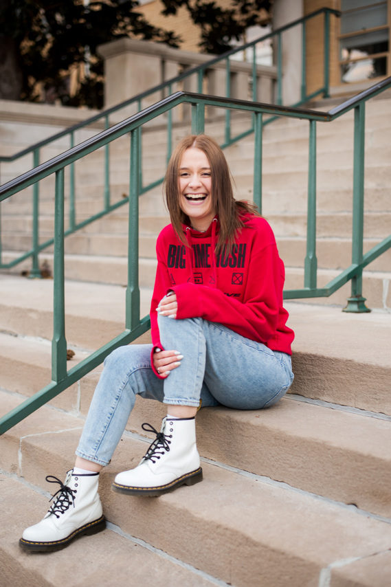 red haired high school senior girl wearing a red hoodie and jeans with white doc martens sits on steps holding one knee and laughing on campus at USAO in Chickasha, Oklahoma