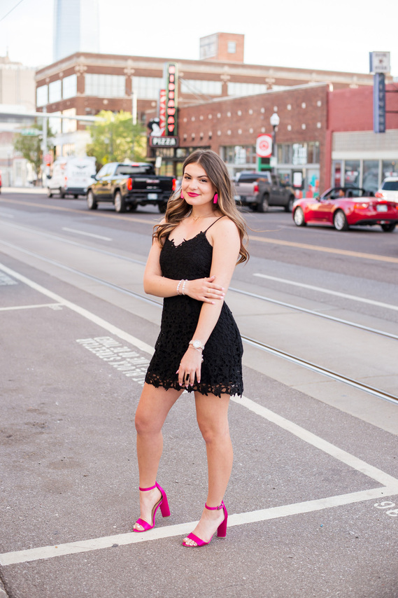 brunette high school senior girl wearing a black dress and pink heels stands on a street with one arm holding the other and smiling in midtown Oklahoma City