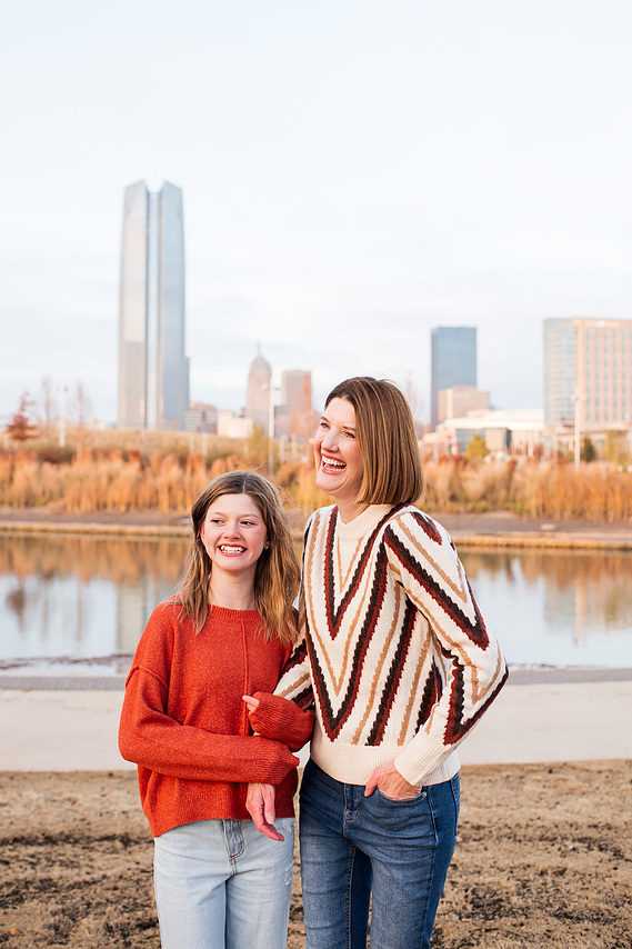 a mom and teen daughter hug each other and smile standing on the lawn of Scissortail Park with downtown Oklahoma City buildings behind them