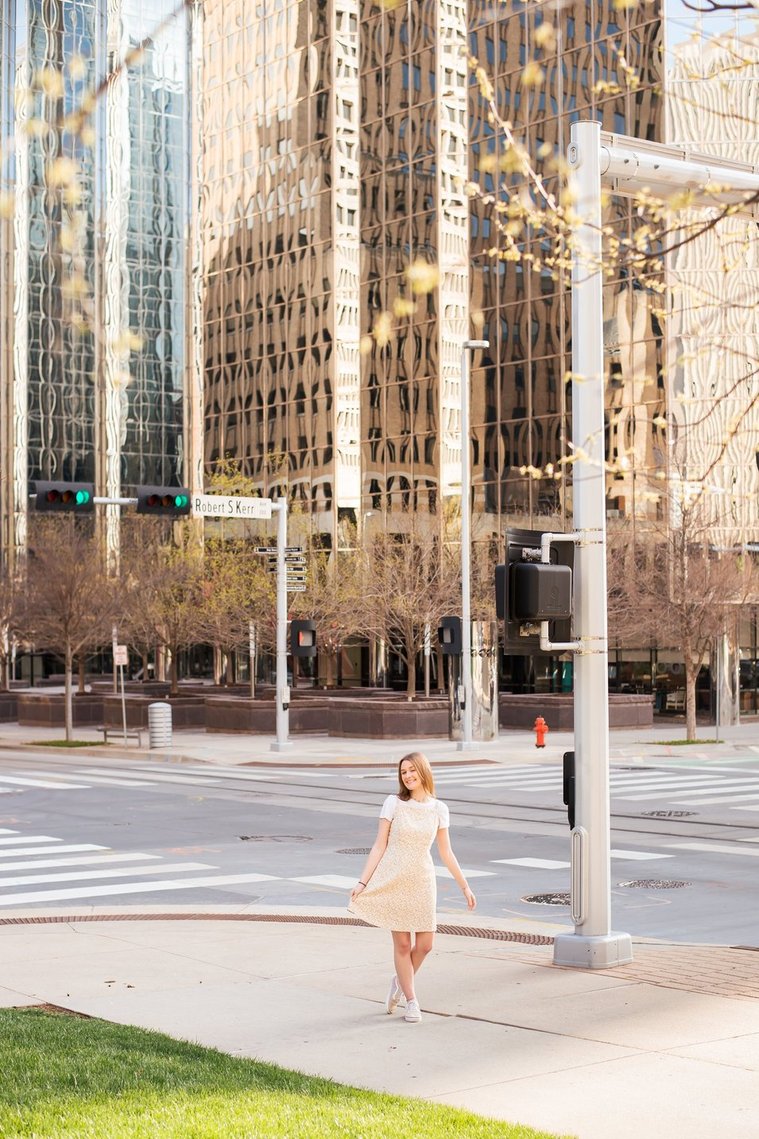 vertical wide angle shot of a high school senior girl standing on the corner of two streets on the sidewalk in downtown Oklahoma City and smiling