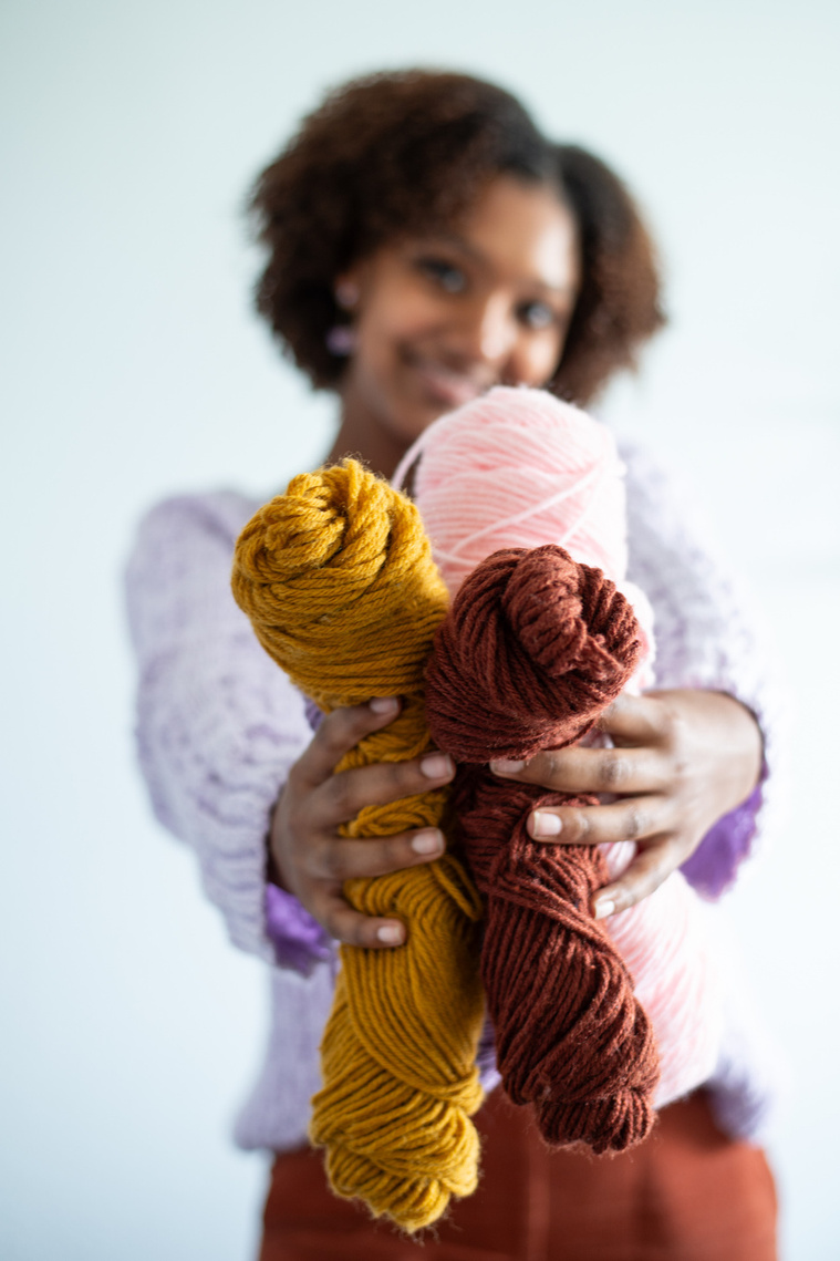 young black girl in a crocheted purple sweater smiles and holds up a handful of yellow, pink and brown yarn close to the camera at a personal brand shoot in Oklahoma