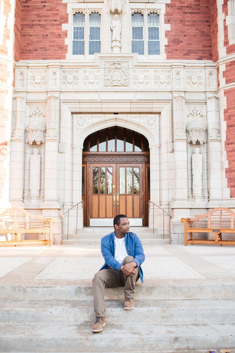 wide angle portrait of an african-american high school senior boy wearing a blue jacket and khakis sitting on the front steps of Evans Hall looking to the side on the OU campus in Norman Oklahoma