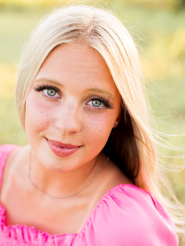 high school senior girl with blond hair wearing a bright pink top looks directly into camera and smiles softly in a field in Oklahoma