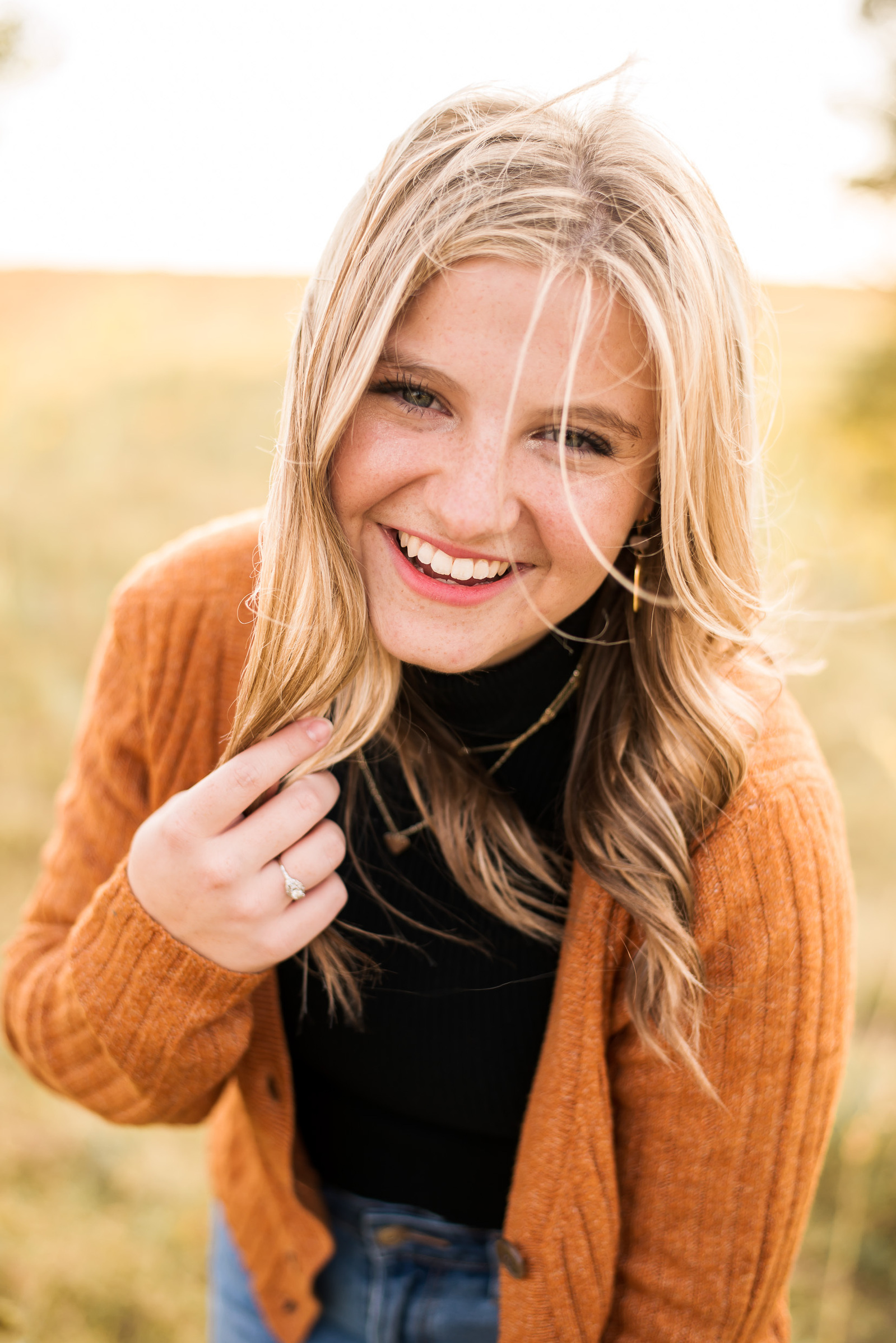 a high school senior girl with long blond hair is leaning toward the camera and smiling wearing an orange cardigan and black turtleneck in a field in Oklahoma