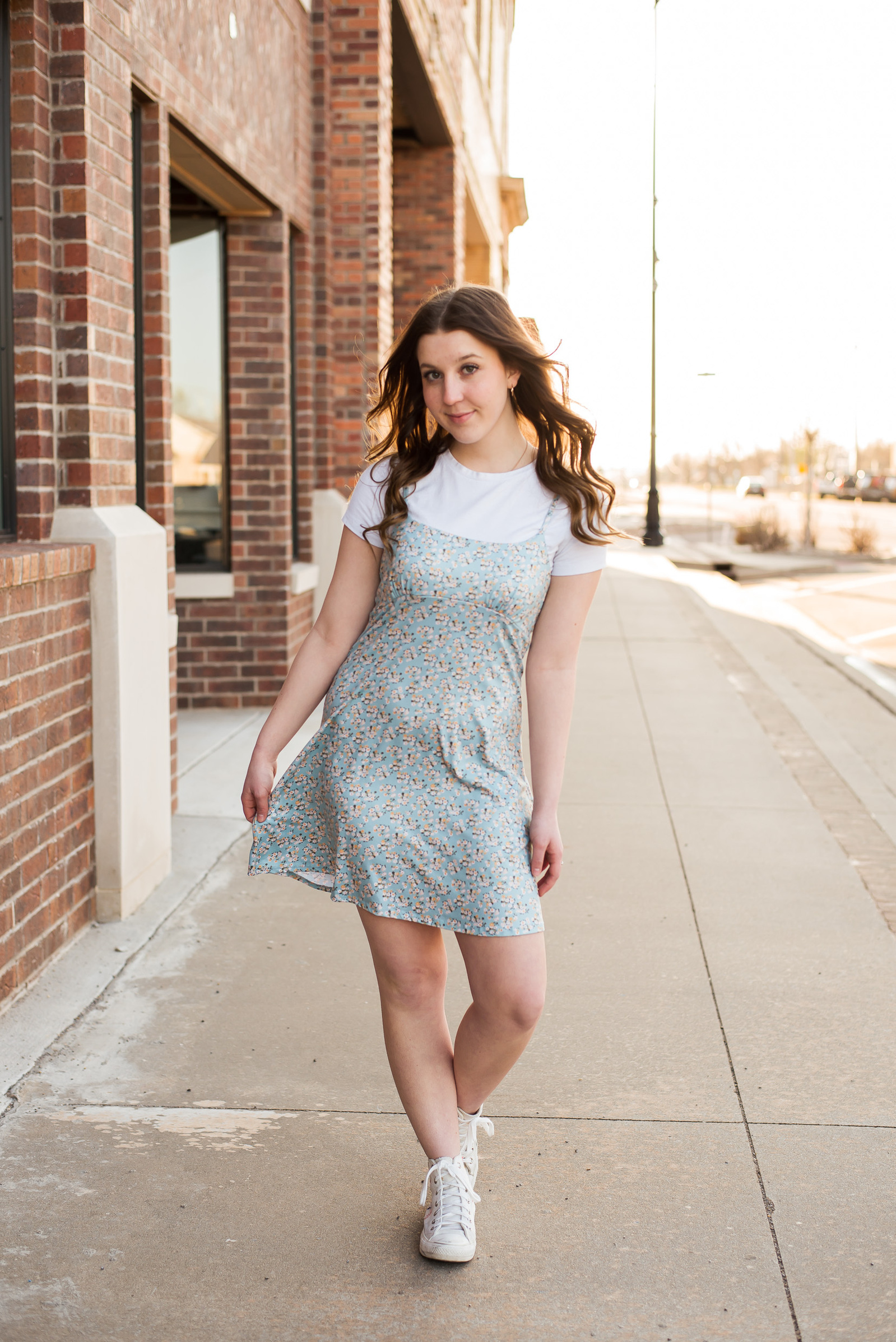 senior girl wearing a light blue sundress with white tee underneath and sneakers walks on a downtown sidewalk and smiles in a small town in Oklahoma