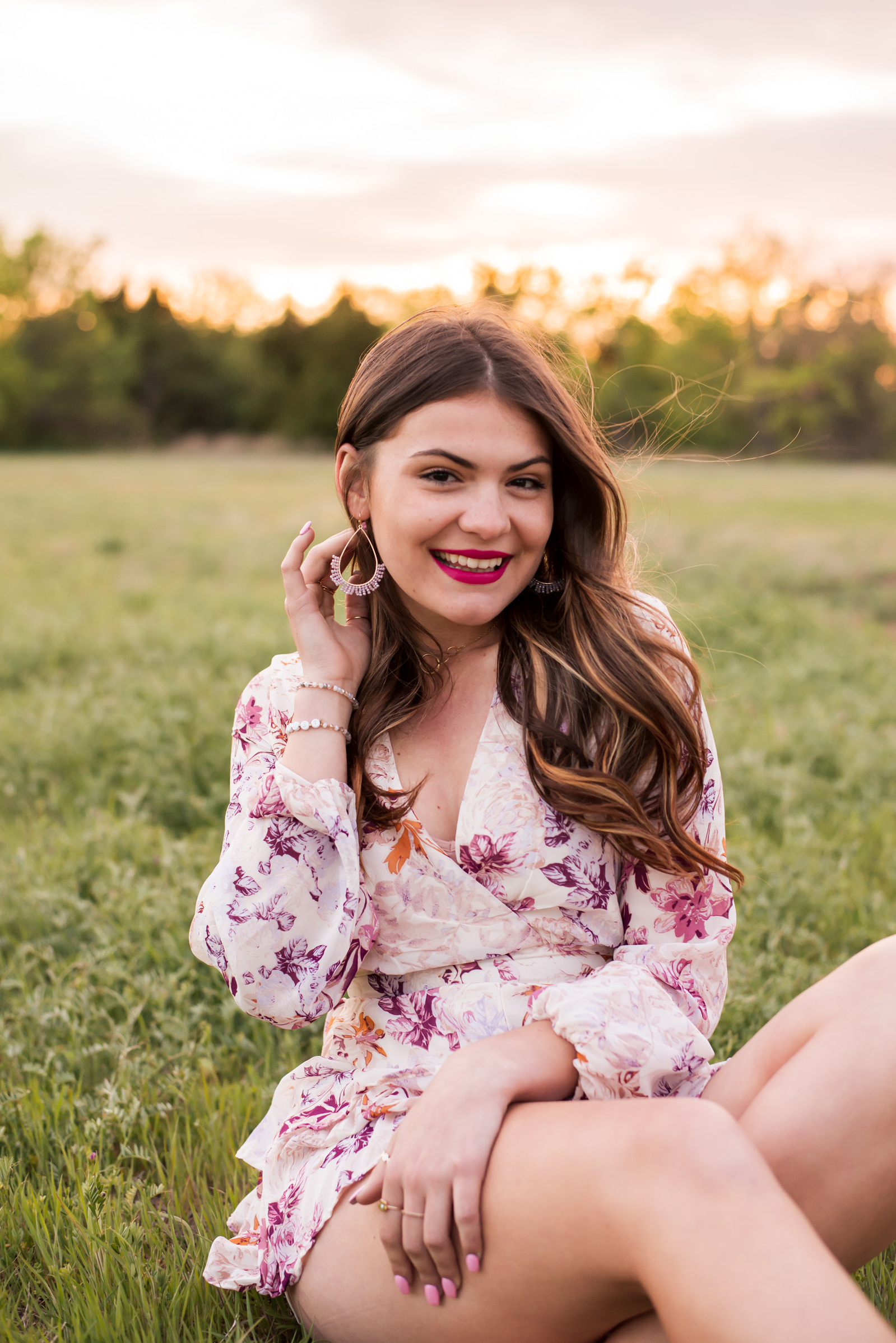 high school senior girl wearing a floral short romper sits and smiles in a field with green grass and the sunset behind her in Oklahoma City