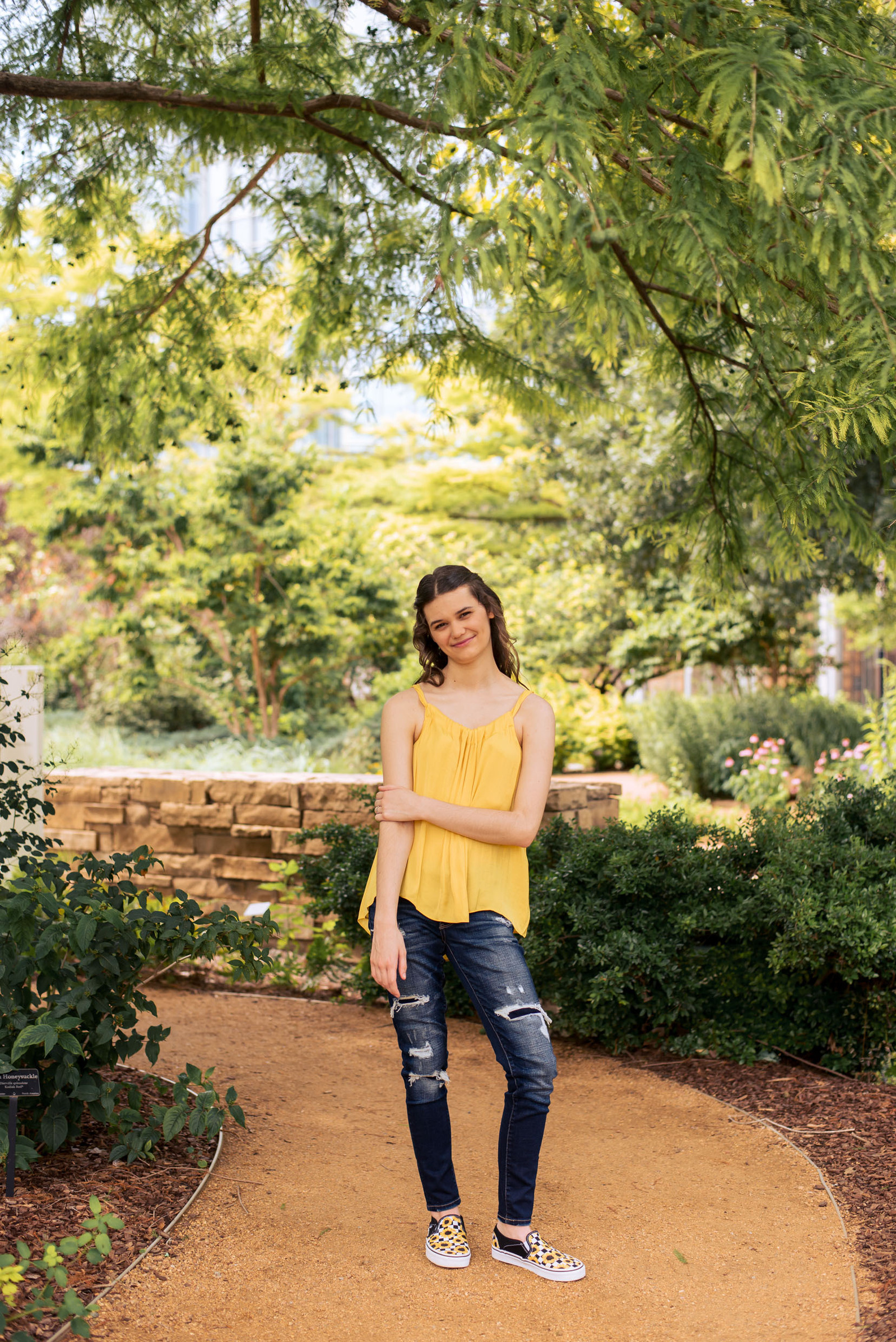 high school senior girl wearing a yellow flowy top and blue jeans with Vans stands on a path with green leaves around her at Myriad Gardens in Oklahoma City