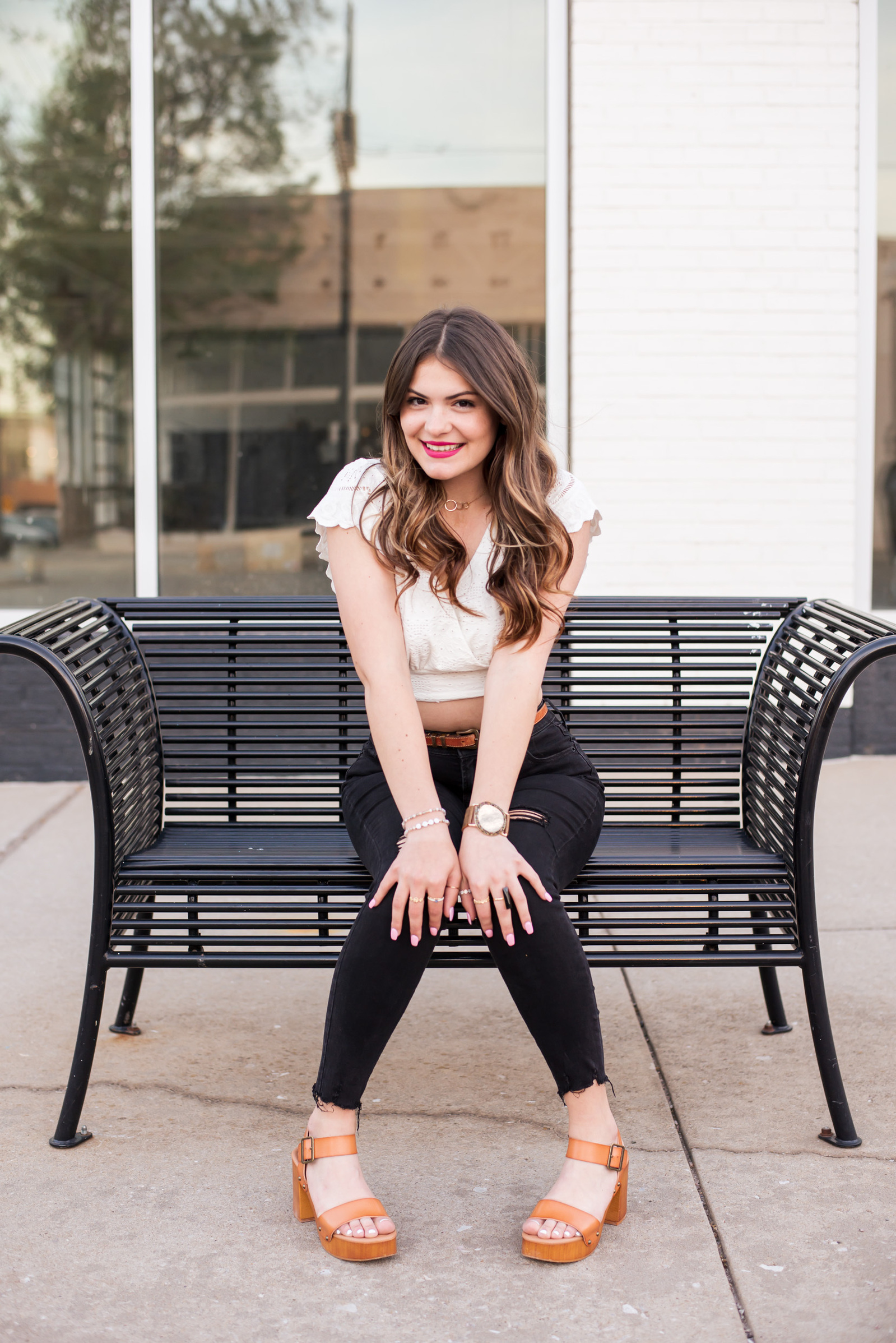 senior girl wearing black pants and a cropped white top sits on a black bench with hands on knees and smiles in Automobile Alley in Oklahoma City