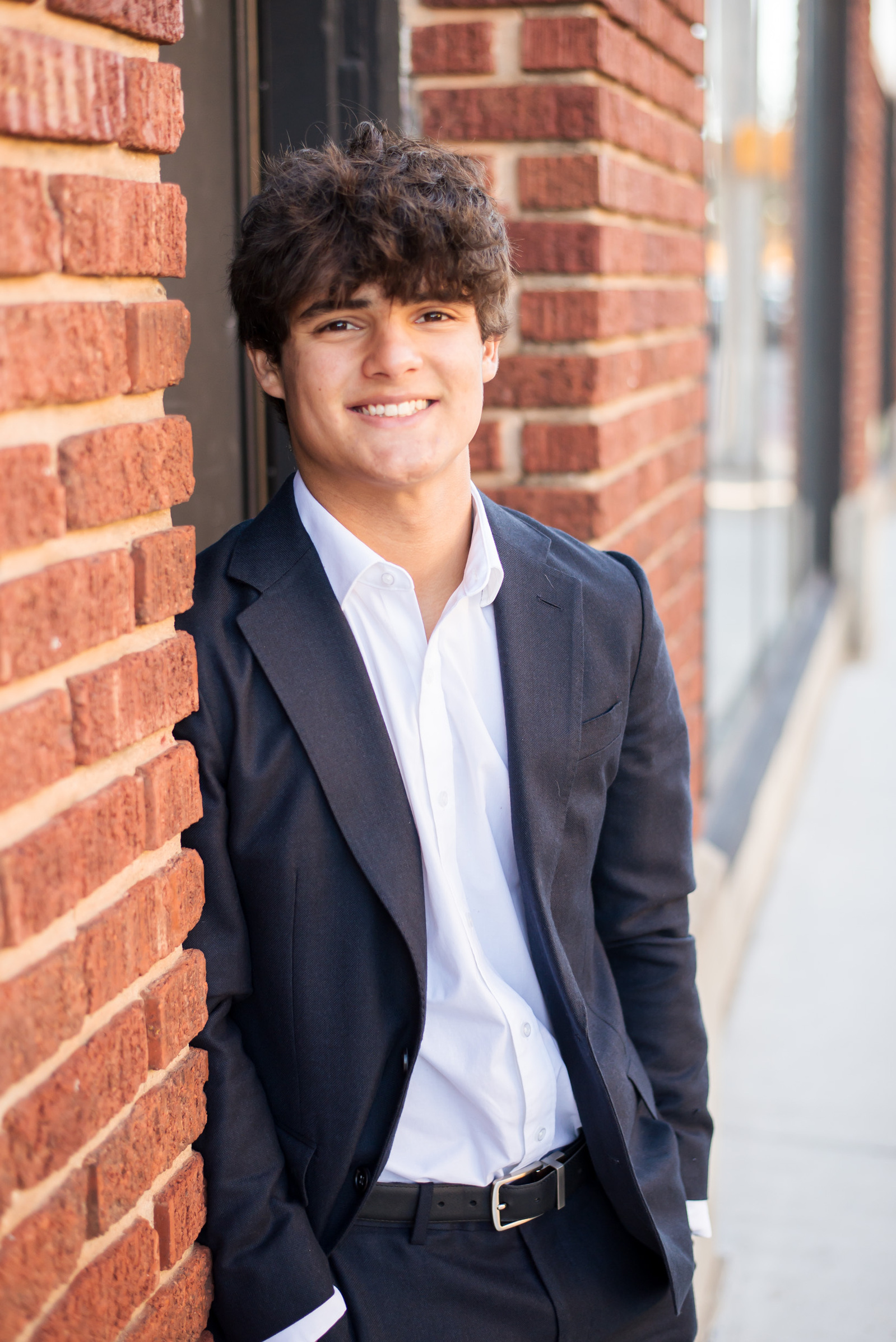 hispanic high school senior boy wearing a dark suit with white shirt leans against a red brick wall with hands in pockets and smiling in midtown Oklahoma City