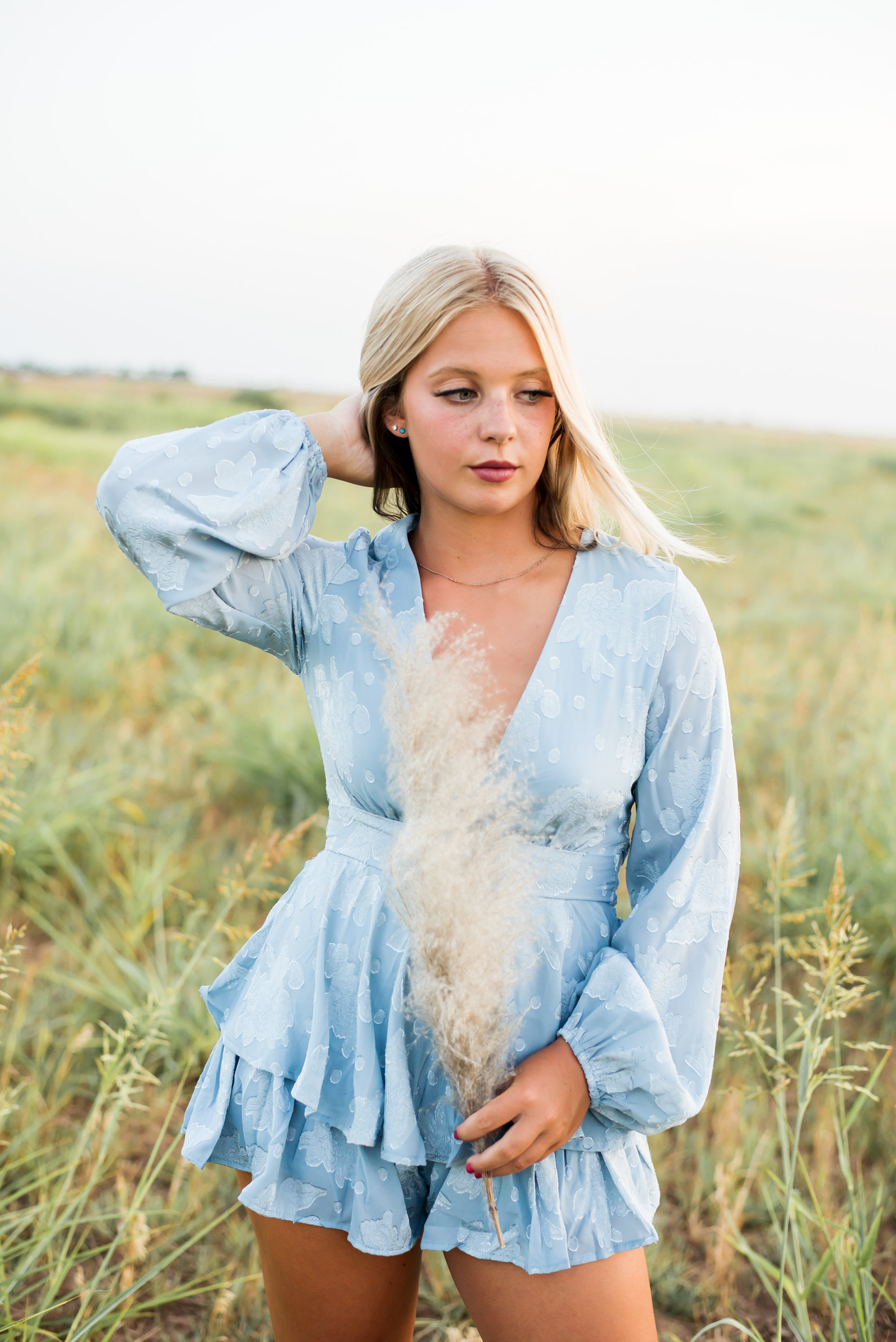 senior girl wearing a blue romper holds a piece of pampa grass and looks to the side with blonde hair blowing standing in a field taken by Oklahoma City senior photographer