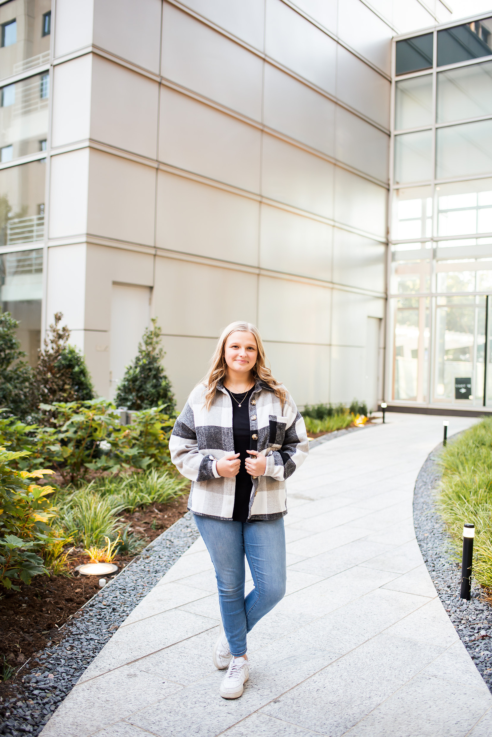 senior girl with blond hair wearing a plaid jacket and jeans stands casually on a sidewalk and smiles in between buildings in downtown Oklahoma City