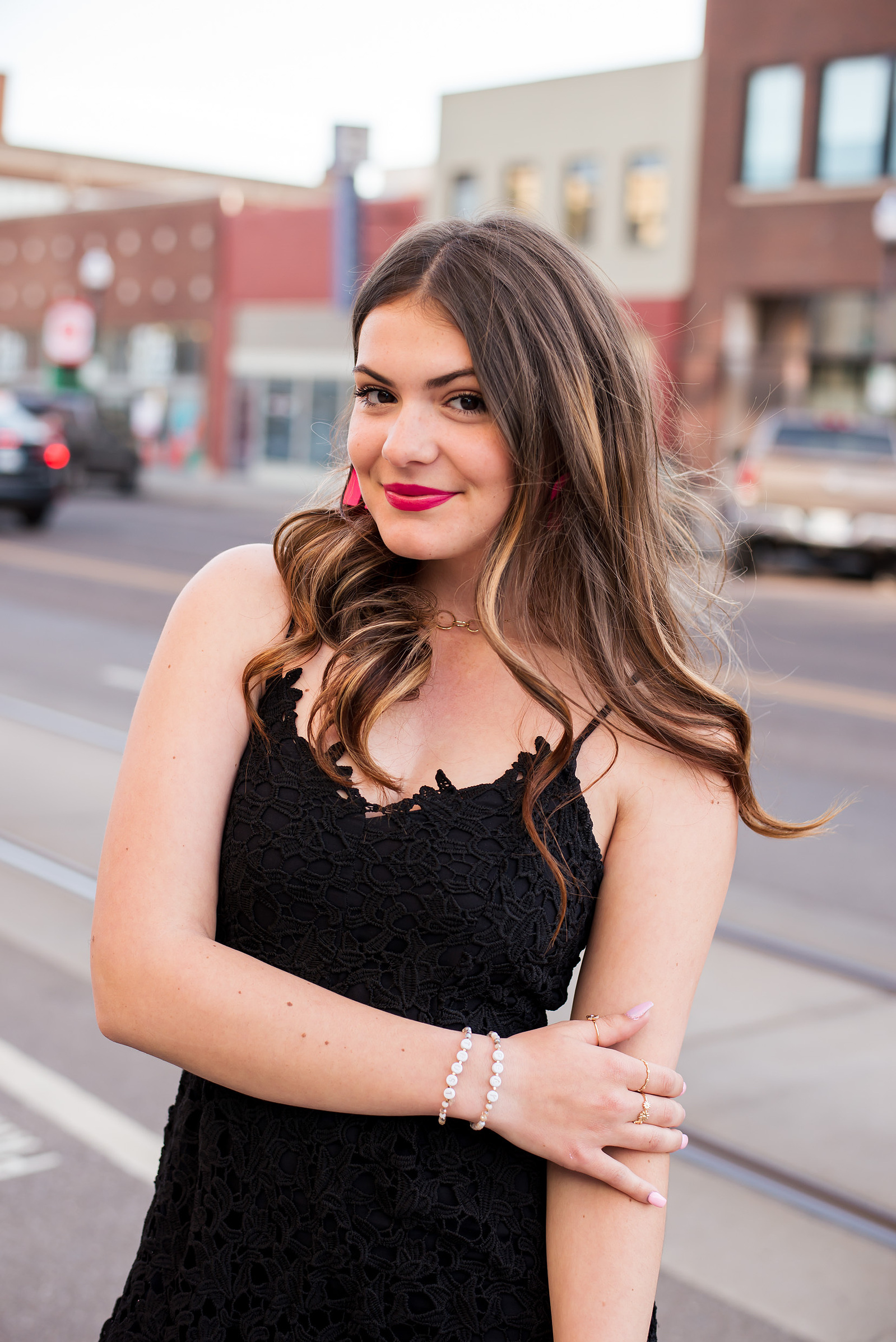 senior girl wearing a black dress with long brown hair holds one arm and smiles cutely while standing near Broadway street in Midtown Oklahoma City