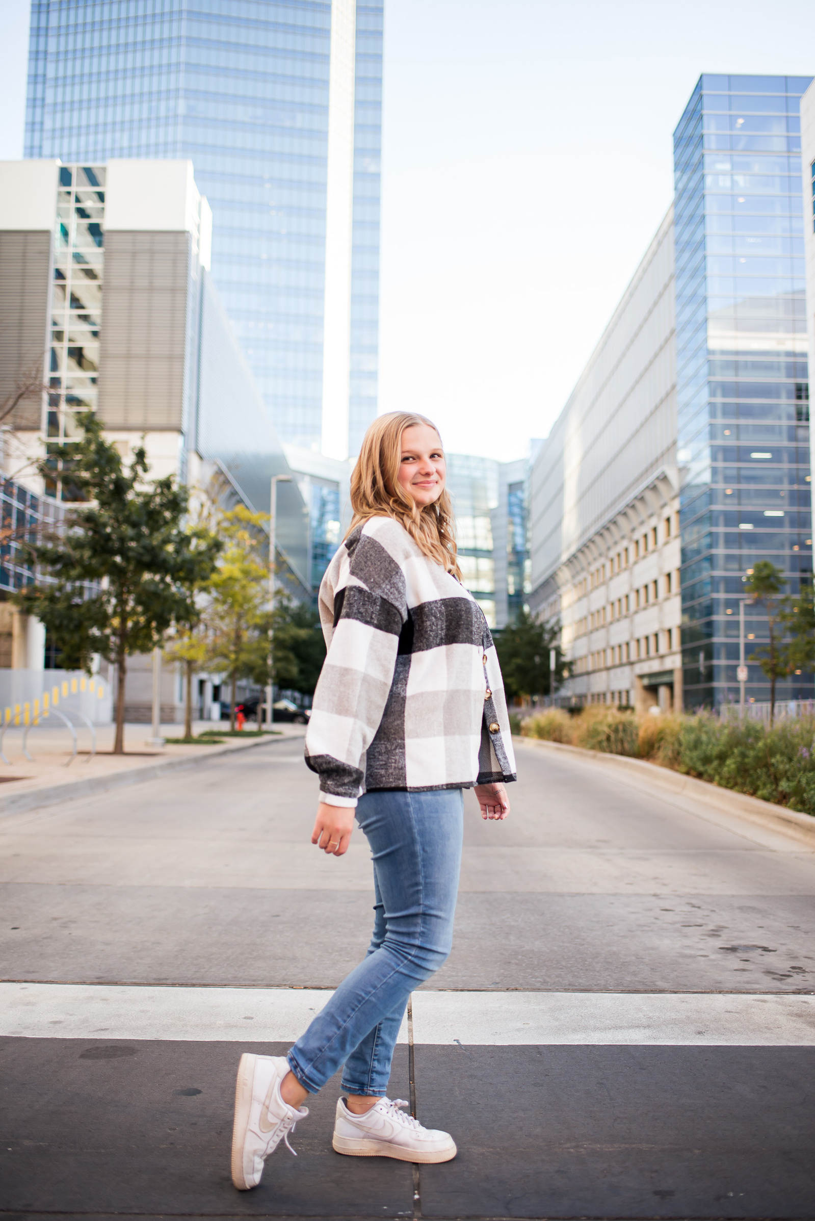 a high school senior girl with blond hair wearing a checkered jacket and blue jeans walks across a street in downtown Oklahoma City with the Devon tower behind her