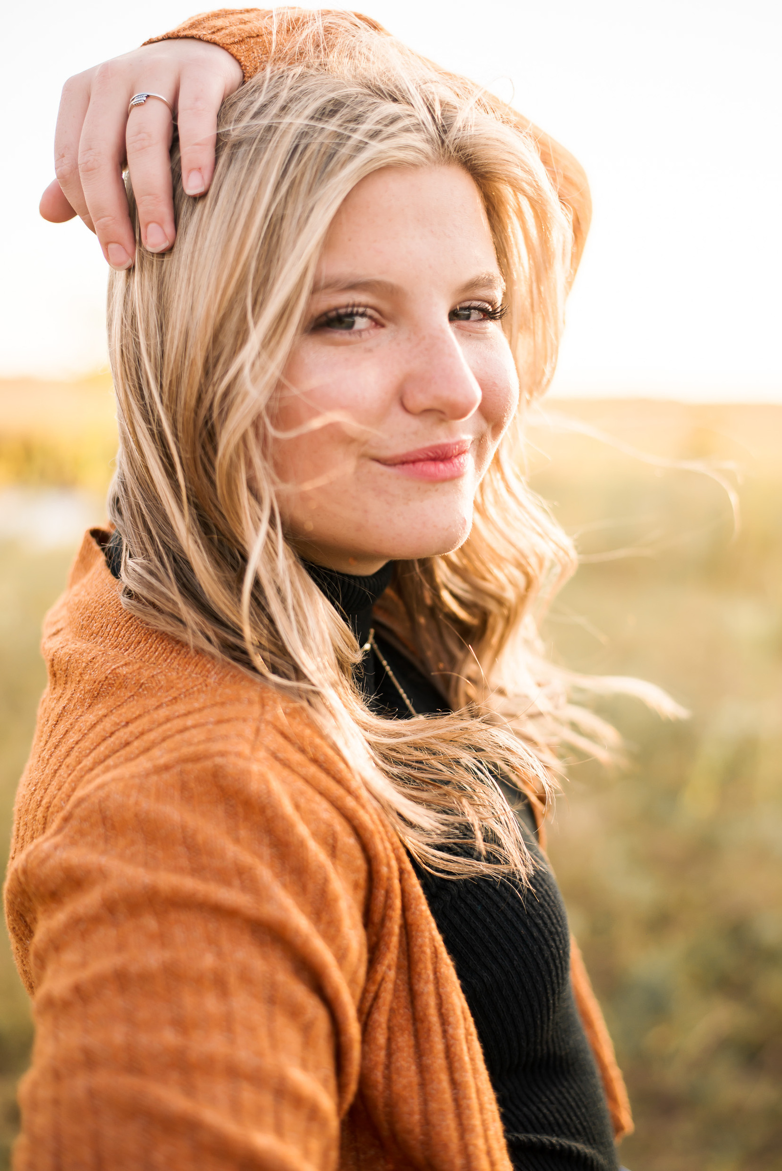 high school senior girl with blond hair wearing an orange sweater stands sideways and looks at camera with a cute smile while her hand rests on her head and the wind blows her hair in a field in Oklahoma