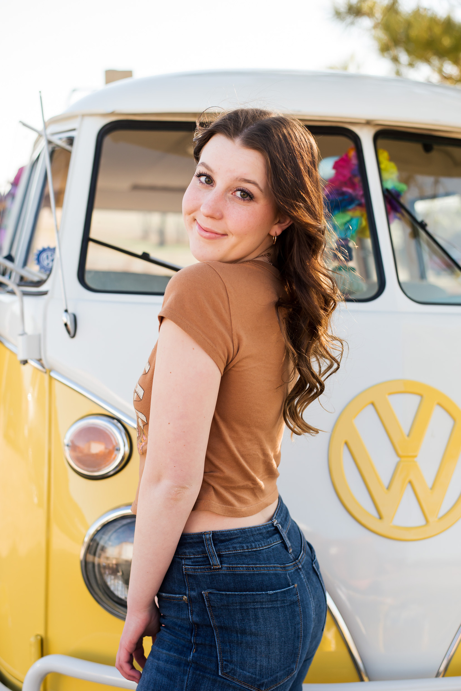 high school senior girl with long brown hair looks over her shoulder and smiles standing in front of a yellow and white Volkswagen bus in Tuttle Oklahoma