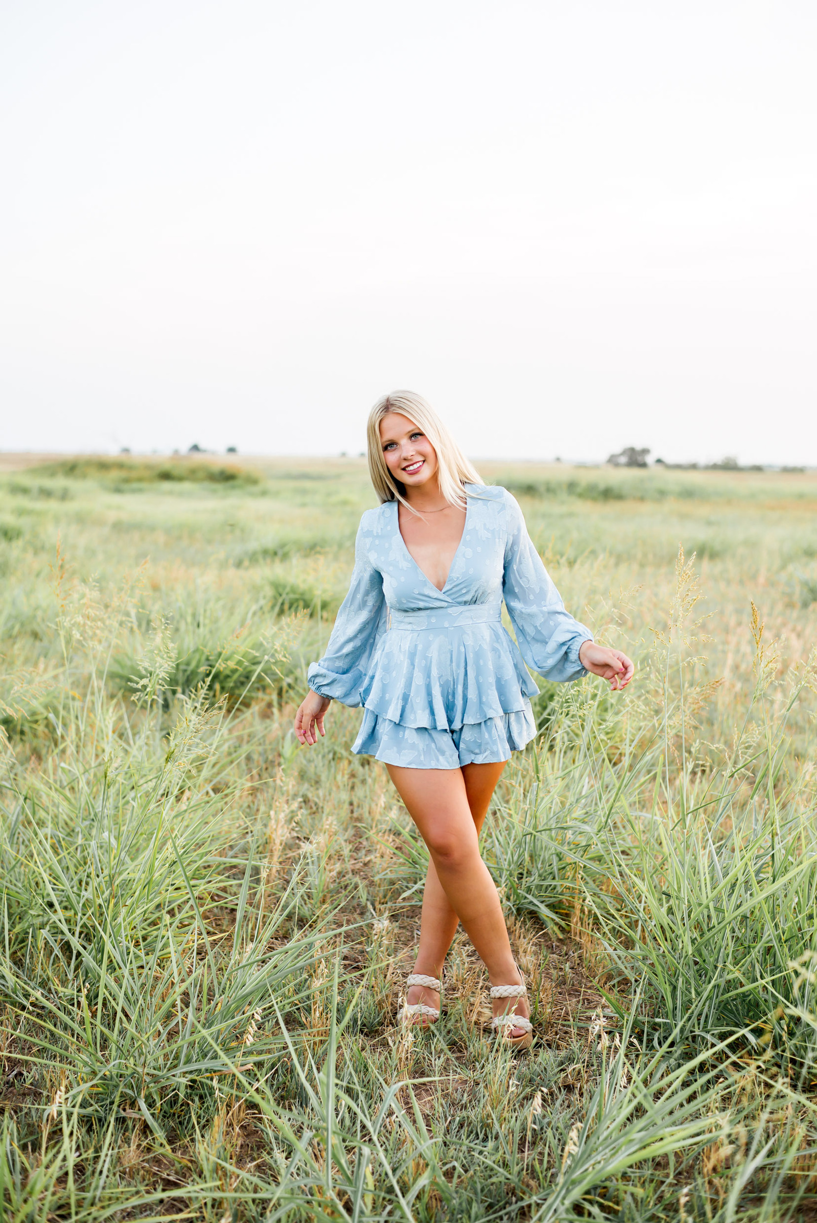 high school senior girl wearing a light blue shorts romper stands crossed at the ankles in the middle of a field in rural Oklahoma