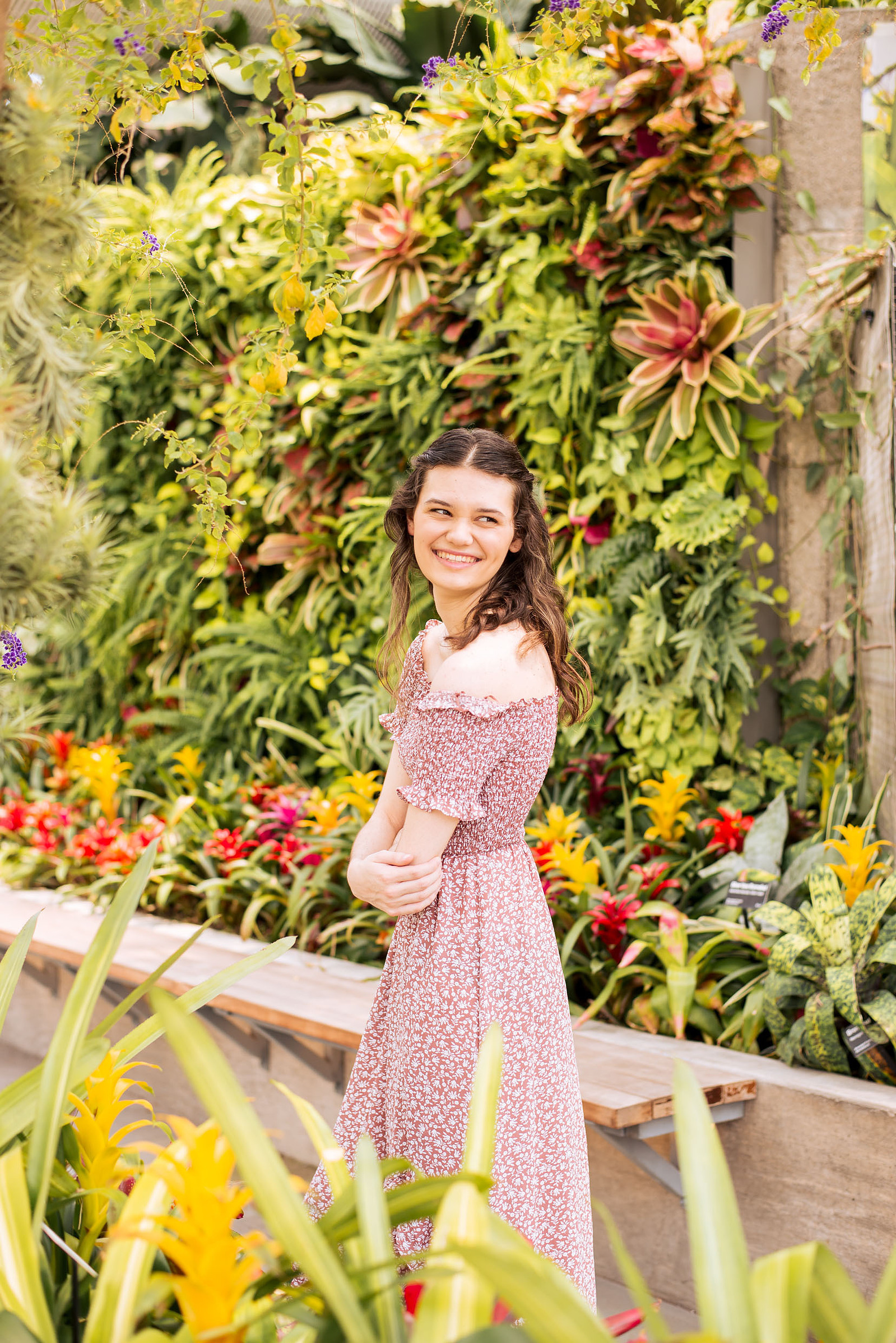 senior girl with brown hair wearing a red print dress looks back over her shoulder smiling with a tropical flower wall behind her inside Crystal Bridge at Myriad Gardens in downtown Oklahoma City