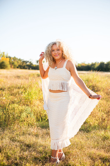senior girl with curly blond hair wearing a matching white top and skirt with fringe holding a white crochet blanket behind her stands and smiles in a field at Martin Nature Park in Oklahoma City