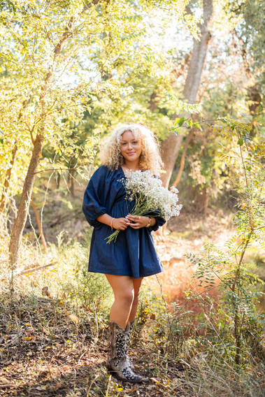 senior girl with curly blond hair wearing a blue dress and cowboy boots holding a bouquet of flowers stands among trees and a creek behind her at Martin Nature Park in Oklahoma City