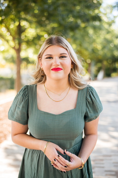 a high school senior girl with long blond  hair and red lips wearing a green dress stands and smiles cutely with hands loosely clasped at waist on a sidewalk at Myriad Gardens in downtown Oklahoma City