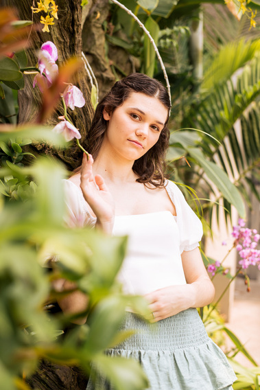 senior girl with brown hair wearing a white top and green skirt leans against a floral wall with foliage in foreground as she looks off softly at Crystal Bridge Conservatory in Myriad Gardens Oklahoma City
