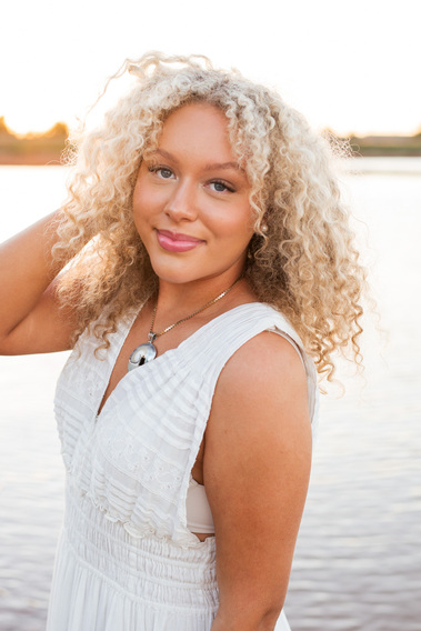 senior girl with curly blond hair wearing a long white dress smiles softly with one hand in her hair with Lake Hefner in Oklahoma City behind her