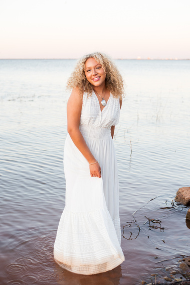 senior girl with curly blond hair wearing a long white dress stands ankle deep in Lake Hefner in Oklahoma City and smiles as she swishes her dress with her hand