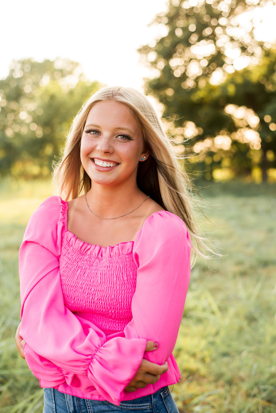 Blond high school senior girl with blue eyes wearing a bright pink top smiles in a field at her senior photo session in Oklahoma.