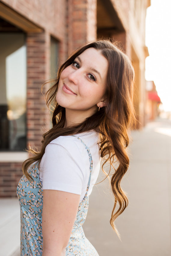 high school senior girl with long brown hair stands on a sidewalk in a small downtown turned to the camera and smiling over her shoulder