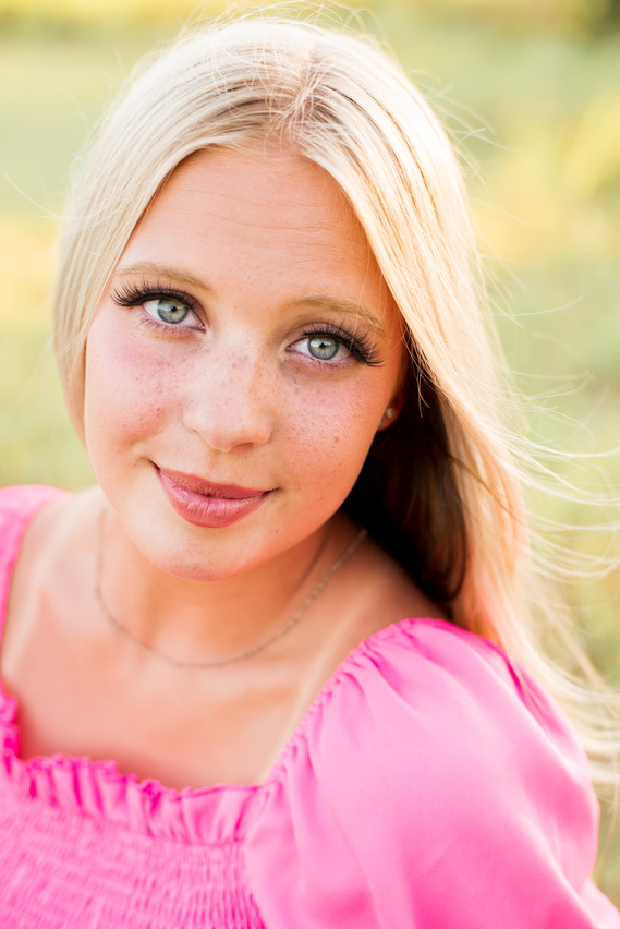close up photo of a blond high school senior girl with big blue eyes wearing a hot pink top looking at camera with a soft smile